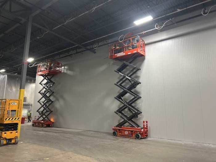 Workers on scissor lifts installing insulated metal wall panels inside a large warehouse under construction.