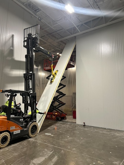 Forklift and scissor lift positioning an insulated metal wall panel during warehouse interior construction.