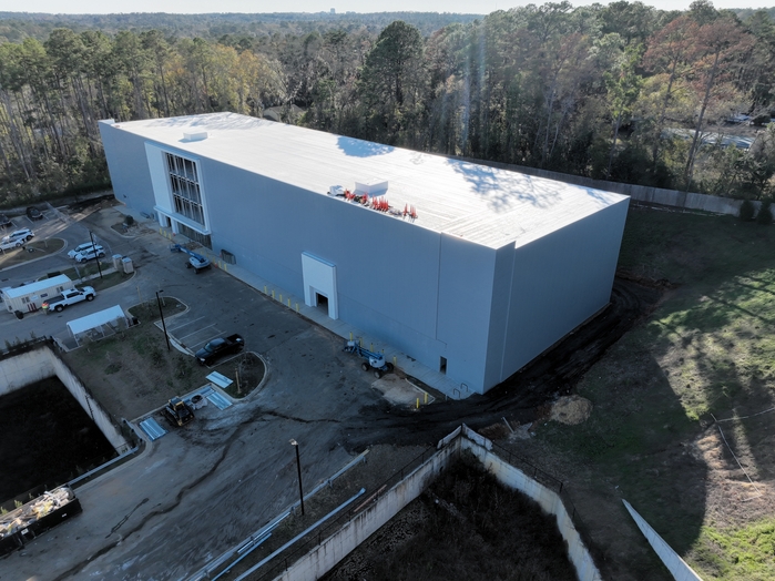 Aerial view of a large commercial warehouse construction site with white exterior panels, parking area, and wooded surroundings.