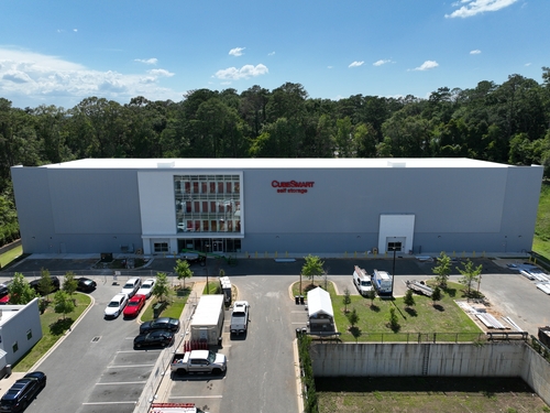 Aerial view of a large commercial self-storage building with parking areas, service vehicles, and surrounding trees.